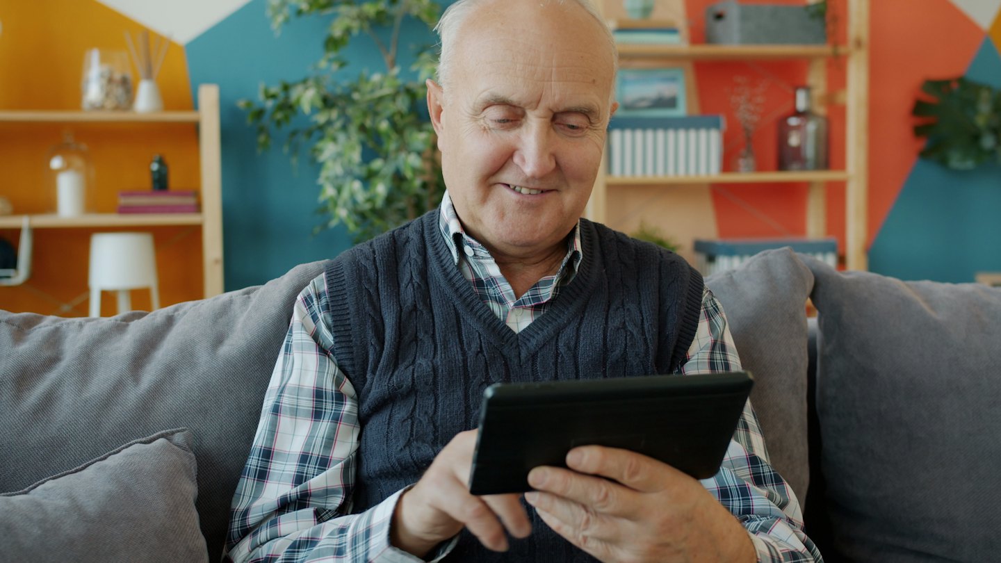 Oudere man glimlacht terwijl hij een digitale tablet gebruikt op een bank.